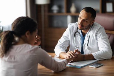 Middle eastern man doctor consoling upset crying sick african american woman patient, therapist supporting sad client, holding hand, giving recommendations, clinic interior. Doctor etiquette concept