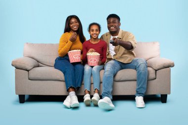 Relaxing together. Happy african american family watching television, parents sitting with their daughter on comfortable sofa over blue background and laughing at the program