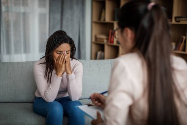 Sad crying black millennial female patient talking with european doctor, need therapy in clinic interior. Consultation with professional, mental disease treatment and medical support, health care