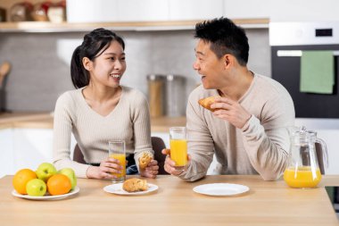 Cheerful korean lovers having snack together at home, happy middle aged man and young woman sitting at kitchen table, eating homemade pastry, drinking fresh orange juice, chatting