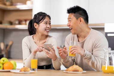 Cheerful korean spouses having breakfast together at kitchen, drinking fresh orange juice, eating pastry, having conversation, checking smartphone, planning weekend, copy space