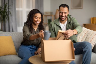 Happy African American Couple Unpacking Cardboard Box After Successful Online Shopping Sitting On Sofa At Home. Spouses Looking At Delivered Items On Weekend. Delivery Service Concept