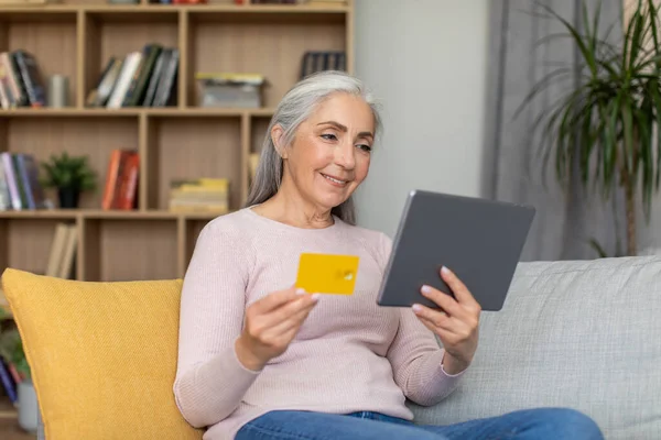 Smiling caucasian old gray-haired female looks at tablet and show credit card in room interior. Online shopping at home, order remotely, finance and banking with technology due covid-19 quarantine