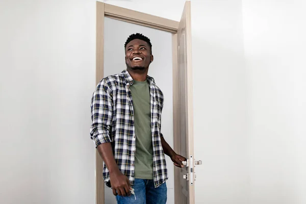 Cheerful Black Millennial Guy Entering His New Rented Apartment Posing Opening Entry Door Looking Aside At Home. Real Estate Offer, Ownership And Purchase Concept