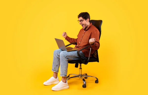 Side View Of Joyful Man Using Laptop And Shaking Fists In Joy Shouting Sitting In Office Chair On Yellow Studio Background. Guy Gesturing Yes Working Online Celebrating Business Success
