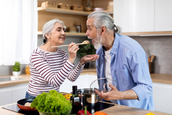 Cheerful senior couple having fun while preparing lunch together in kitchen, loving senior woman feeding her husband with spaghetti, giving him a try while cooking food at home, closeup