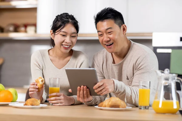 Cheerful asian couple using digital tablet while having breakfast at kitchen, loving man and woman eating healthy food, checking photos on social media and smiling, having video conference with family