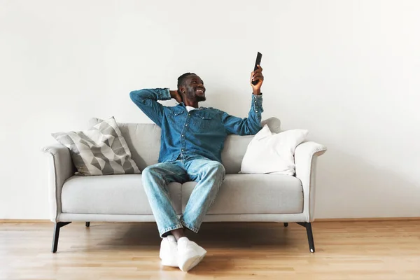Happy African American Man Turning On AC Conrolling Cooling And Heating With Air Conditioning System Pointing Remote Controller Up Sitting On Sofa Relaxing At Home. Climate Control For House