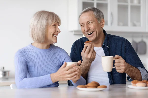 Happy beautiful elderly spouses drinking coffee with fresh pastry at home, sitting at kitchen table, holding mugs, having conversation and smiling, enjoying time together, closeup