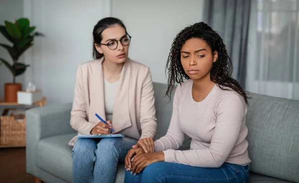 Mental medical support. Sad young european lady doctor in glasses calms black woman patient in office clinic interior. Session with professional, help from depression, psychotherapy and health care