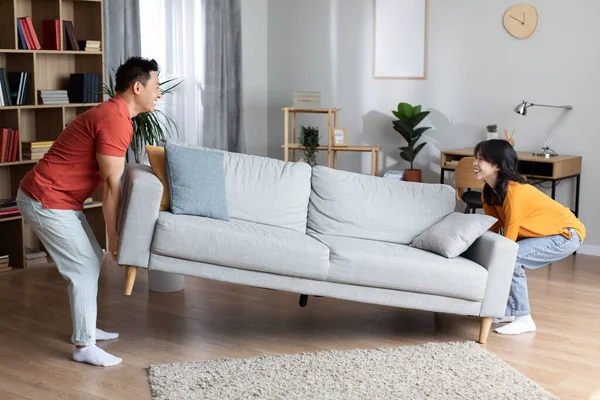 Happy asian family moving furniture at their new house, positive middle aged husband and young wife pushing sofa in cozy living room, enjoying new apartment, full length, copy space