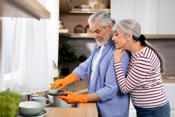 Loving Senior Woman Embracing Her Husband From The Back While He Washing Dishes In Kitchen, Happy Elderly Spouses Sharing Domestic Chores, Caring Husband Helping With Cleaning, Side View