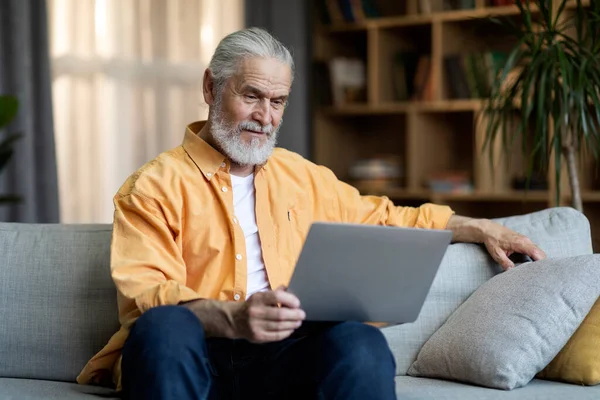 Happy long-haired bearded senior man in casual outfit sitting on couch, using modern laptop, reading content or email, spending time alone at home, living room interior, copy space