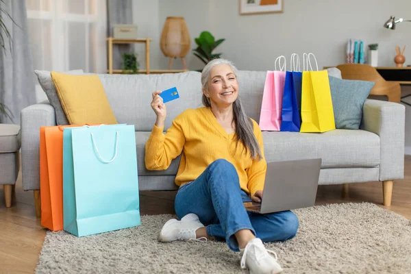 Satisfied caucasian old gray-haired female sits on floor with laptop and a lot of packages with purchases, show credit card in living room interior. Online shopping, shopaholic at home and big sale