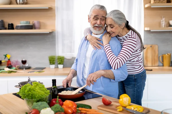 Loving Wife Embracing Her Elderly Husband While He Cooking Lunch In Kitchen, Happy Senior Spouses Preparing Tasty Food And Bonding At Home, Enjoying Spending Retirement Time Together, Copy Space