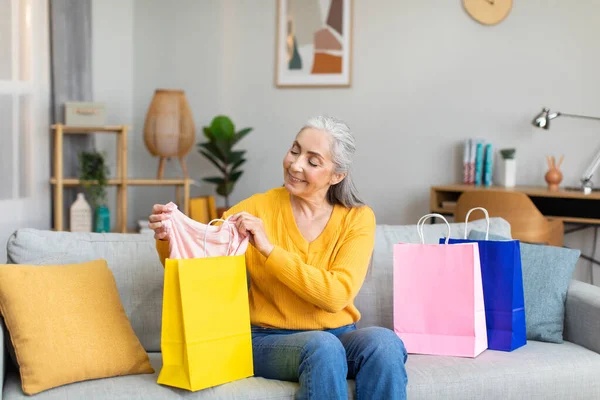 Smiling elderly european gray-haired lady unpacks package with new clothes on sofa in living room interior. Modern shopping in store, huge sale, great offer at home, ad and order delivery due covid-19