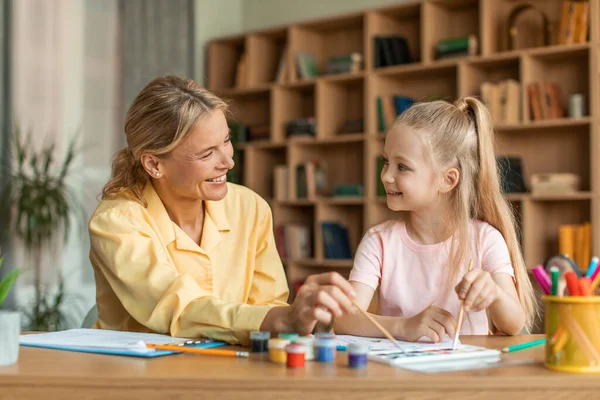 Pretty little girl painting with child specialist, sitting at table with female teacher and holding brush, female teacher doing arts and crafts with child during private lesson, copy space