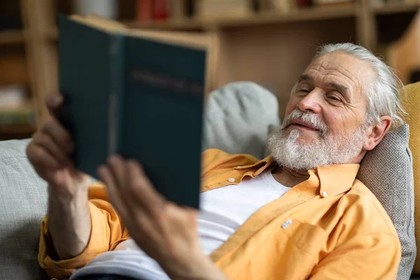 Relaxed attractive grandfather chilling on couch at home, positive bearded senior man lying on sofa in living room, reading book and smiling, enjoying story, closeup shot