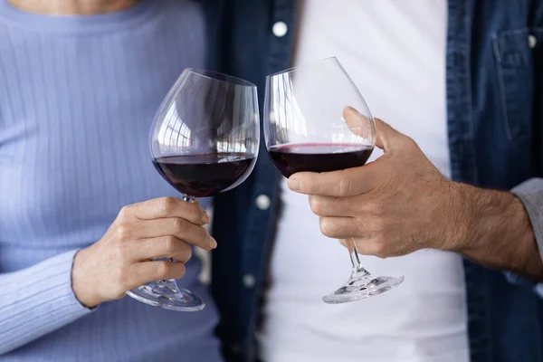 Cropped of couple drinking red wine at kitchen, unrecognizable man and woman in casual outfits hugging and cuddling, holding glasses with drink, enjoying time together, closeup