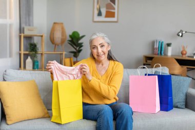 Cheerful caucasian senior gray-haired lady shows clothes from package, sits on sofa after shopping in room interior. Shopaholic enjoy purchases at home. Sale, fashion unpacking blog and lifestyle
