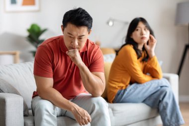 Selective focus on pensive middle aged asian man sitting on couch at home, having fight with young wife, upset korean lady looking at her thoughtful lover, suffering from crisis in marriage