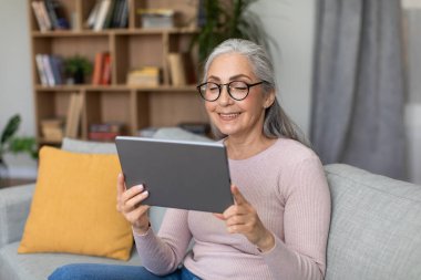 Happy caucasian aged gray-haired lady in glasses chatting on tablet, watch video lesson in living room interior. Modern tech, work remotely, meeting and video call at home during covid-19 pandemic