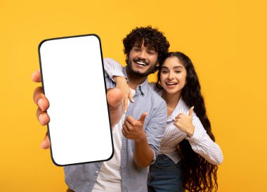 Cheerful Indian Couple Demonstrating Big Blank Smartphone And Showing Thumbs Up, Happy Man And Woman Recommending New Mobile App Or Website, Standing Isolated On Yellow Background, Mockup, Collage