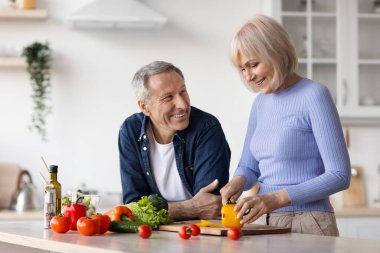 Attractive happy senior woman cooking healthy delicious meal and talking to her handsome husband, attractive elderly spouses enjoying time together at home, white kitchen interior, copy space