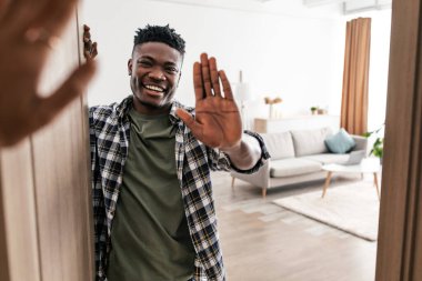 Hello. Cheerful Black Man Waving Hand Opening Door Of His House Standing Indoor. Male Real Estate Owner Gesturing Hi Smiling Looking At Camera Posing In Rented Apartment. Property Ownership