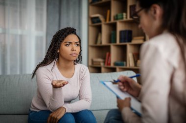 Unhappy sad black millennial woman patient talking with european doctor in clinic interior. Medicine, professional help, depression treatment and support, patient care, therapy and health problems