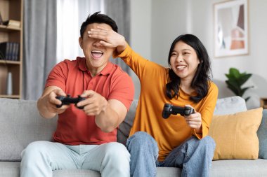 Cute chinese lovers having fun at home, happy asian man and woman sitting on couch, holding joysticks, playing video games, laughing lady covering her boyfriend eyes, cheating in game