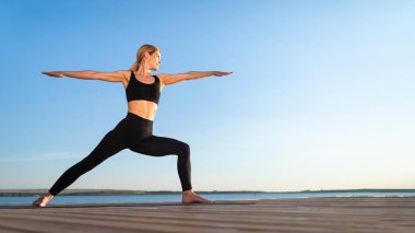 Beautiful Middle Aged Woman Practicing Yoga Outdoors, Standing In Warrior Pose, Portrait Of Fit Female In Activewear Training On Wooden Pier Near Lake, Enjoying Healthy Lifestyle, Panorama