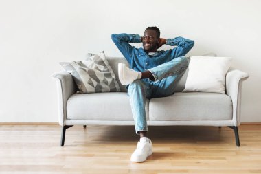 Happy Black Guy Sitting On Sofa Holding Hands Behind Head Relaxing Looking Aside Enjoying Weekend At Home. Millennial Man Resting In Modern Living Room Indoors