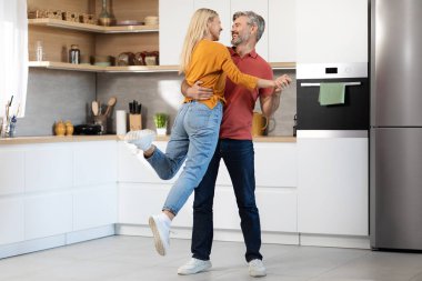 Loving middle aged couple in casual outfits having fun while cooking together at kitchen, dancing, looking at each other and smiling, happy husband lifting up his laughing beautiful wife, copy space