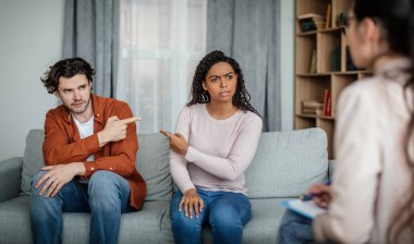 Offended sad millennial black female pointing finger at european man on meeting with psychologist in office clinic interior, panorama. Family psychological therapy, accusation, quarrel and scandal