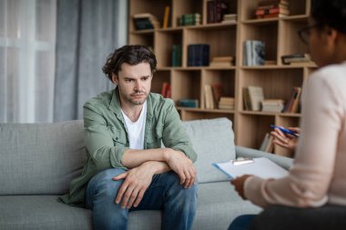 Despaired depressed young caucasian man suffers from mental problems, listens to black female psychologist in clinic interior. Consultation, medical care, psychological treatment and professional help
