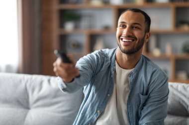 Portrait Of Cheerful Young African American Man Watching TV At Home, Smiling Millennial Black Guy Holding Remote Controller And Switching Channels While Relaxing On Couch In Living Room, Closeup