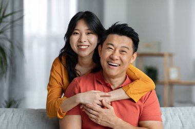 Happy asian man and woman in casual outfits sitting on couch in living room, hugging and smiling, posing together, enjoying weekend at home, having fun, copy space, closeup portrait