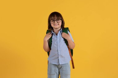 Schooltime concept. Studio portrait of little clever boy preschooler wearing eyeglasses smiling to camera with backpack, yellow background