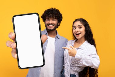 Mobile Offer. Cheerful Indian Couple Pointing At Big Blank Smartphone With White Screen While Standing Over Yellow Background In Studio, Happy Young Man And Woman Advertising New App, Mockup