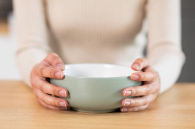 Female hands holding empty dish plate, closeup photo. unrecognizable hungry woman sitting at wooden table with clear bowl, waiting for food, cropped, home interior, copy space