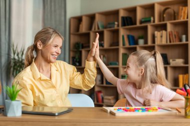 Happy pretty girl giving her female teacher high five, woman and child preschooler sitting at table together, learning alphabet during private lesson and celebrating success