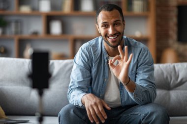 Smiling Young Black Man Using Smartphone On Tripod For Recording Video At Home, African American Male Blogger Capturing Content For Social Networks, Showing Ok Gesture At Camera, Selective Focus