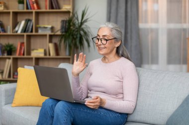 Smiling caucasian senior gray-haired female in glasses waving her hand at laptop webcam in living room interior. Modern device, chat in social networks, meeting remotely and communication due covid-19