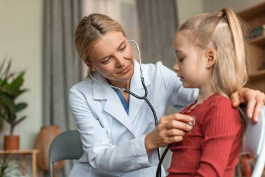 Cheerful female pediatrician holding stethoscope and examining child girl patient, checking heart lungs of kid, doing pediatric checkup in hospital children medical care concept