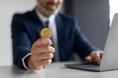 Cryptocurrency Trading. Unrecognizable Smiling Businessman Sitting At Desk With Laptop And Showing Golden Bitcoin, Middle Aged Male Recommending Online Platform For Mining, Selective Focus, Closeup
