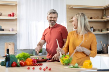 Loving middle aged spouses preparing healthy dinner together, cheerful attractive blonde wife and handsome husband cooking at home, having conversation and smiling, kitchen interior, copy space