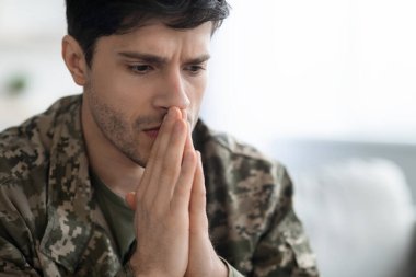 Serious short-haired young man in military uniform soldier looking down, holding palms by mouth, upset guy suffering from posttraumatic syndrome after service, closeup photo, panorama with copy space