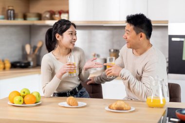 Happy asian spouses having fun while eating at kitchen, laughing and gesturing, cheerful loving husband and wife drinking fresh juice, eating fruits and pastry, having conversation, copy space