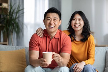 Positive asian middle aged husband and young pretty wife sitting on couch in living room, hugging, watching movie, drinking tea, happy korean man and woman enjoying TV show, copy space